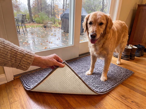 A hand lifts the corner of a grey Muddy Mat to show the cream non slip backing while a golden retriever stands on the mat