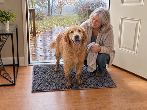 A woman with silver hair crouches beside a golden retriever with muddy paws and legs standing on a grey Muddy Mat inside an open door, a rainy deck outside and the floor clean