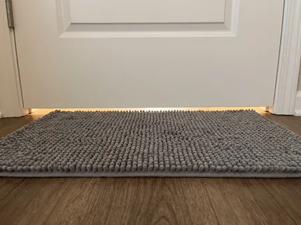 Close view of the bottom edge of a white door with a clear strip of floor light between the door and the top of a thick grey Muddy Mat lying in front of it