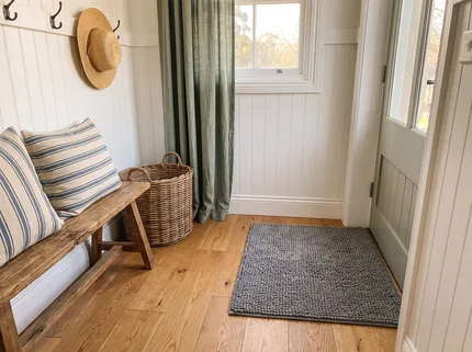 A grey Muddy Mat just inside a side entry with white beadboard walls, a wooden bench with striped cushions and a wicker basket, the oak floor clean around the mat