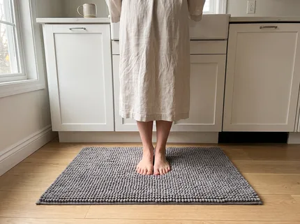 Bare feet standing on a grey Muddy Mat in front of a kitchen sink, a linen robe, clean light wood floor around the mat