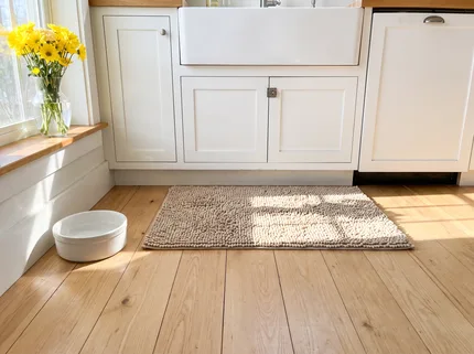 A beige Muddy Mat on the clean wood floor in front of a sunny kitchen sink, a ceramic dog water bowl beside it and yellow flowers on the windowsill