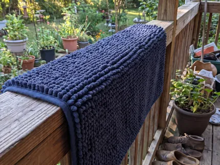 A navy blue Muddy Mat draped over a wooden porch railing to be shaken out, garden plants and shoes behind it