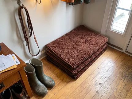 Three chocolate brown Muddy Mats folded and stacked in a back entry beside a pair of rubber boots