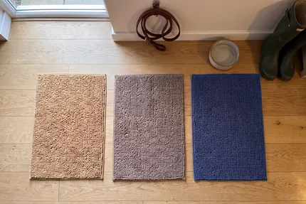 A beige, a grey and a navy blue Muddy Mat laid side by side on a clean oak floor