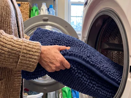 Hands feeding a navy blue Muddy Mat into the open door of a front loading washing machine